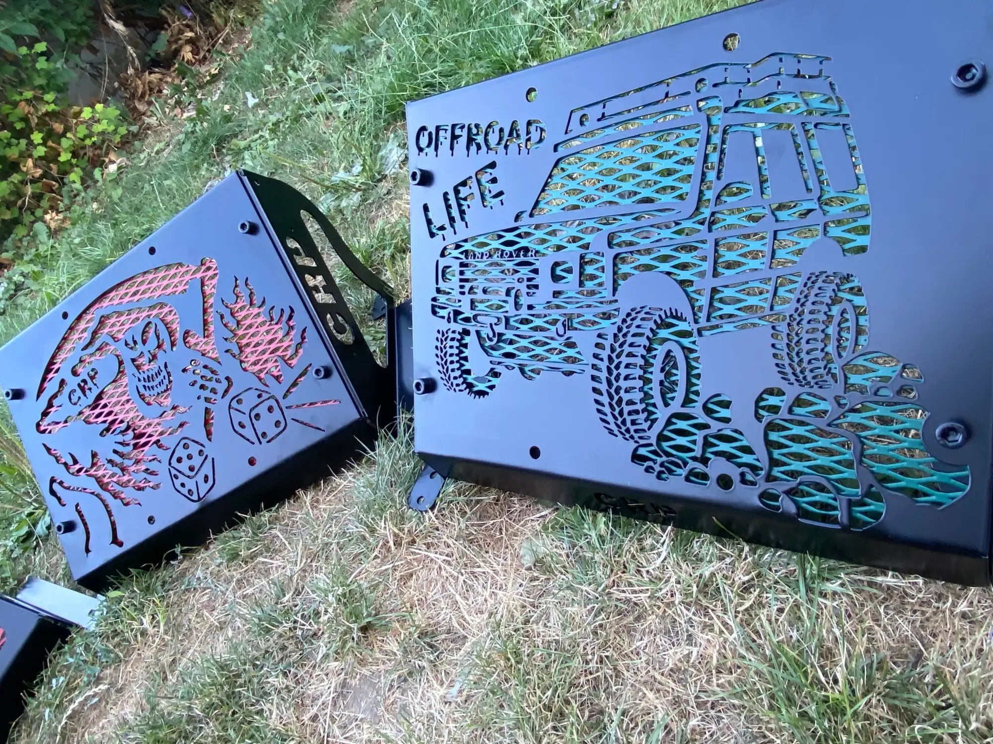 Black metal outdoor grill guard with laser-cut designs featuring a green off-road vehicle and red skull-and-dice motifs.
